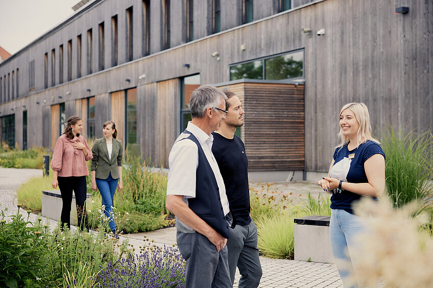 Im Garten des NAWAREUMs spricht eine Museumsführerin mit zwei Besuchern. Im Hintergrund spazieren zwei Frauen und unterhalten sich.