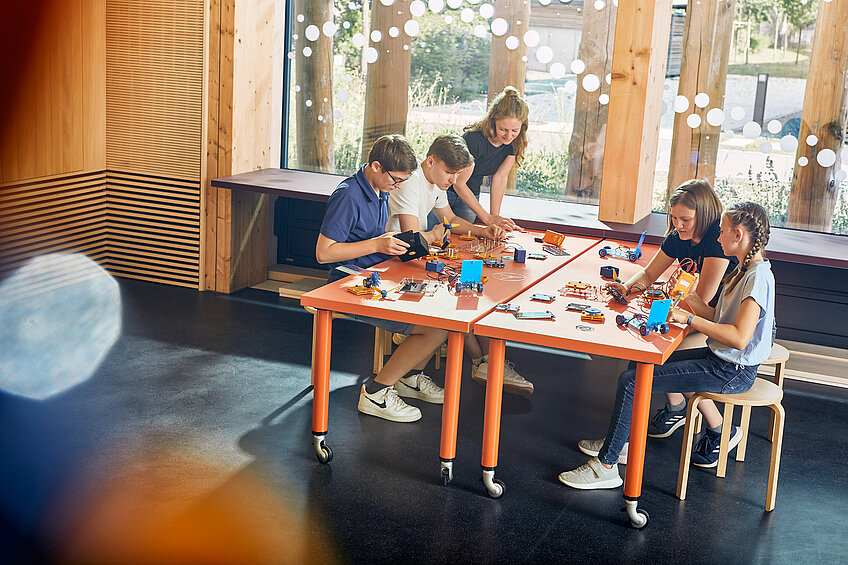 Vier Kinder sitzen an einem großen Tisch und basteln mit Elektronik-Bauteilen. Eine erwachsene Frau hilft ihnen dabei.