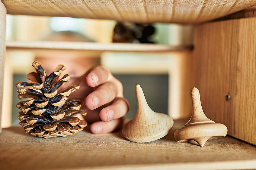 Eine Kinderhand greift nach einem Fichtenzapfen, der in einem Regal liegt. Daneben liegen zwei Holzkreisel.